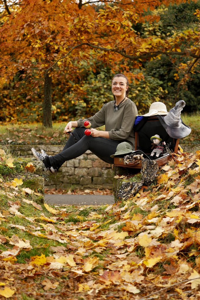 Lächelnde Frau sitzt im Herbstpark neben offenem Koffer mit Hüten und Requisiten.