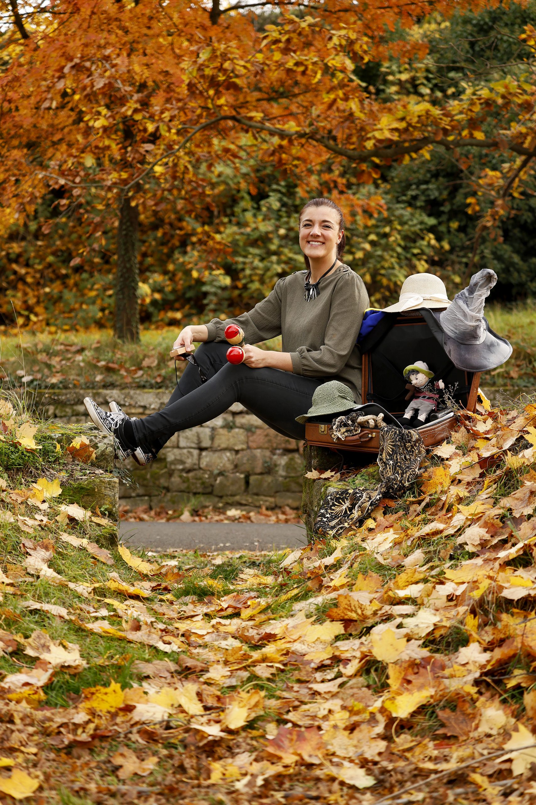 Lächelnde Frau sitzt im Herbstpark neben offenem Koffer mit Hüten und Requisiten.