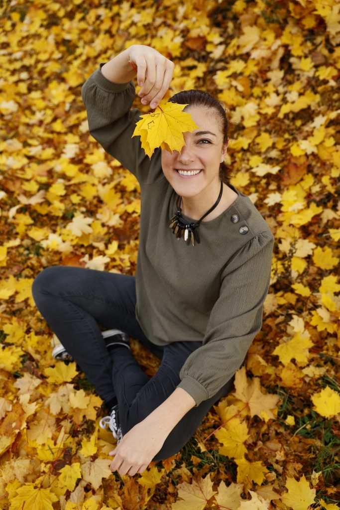 Lächelnde Frau sitzt im Herbstlaub und hält ein gelbes Ahornblatt vor ein Auge.