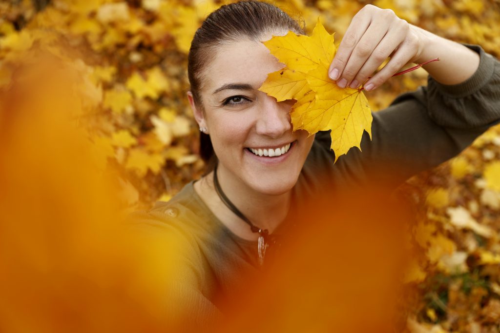 Lächelnde Frau hält ein gelbes Ahornblatt vor ein Auge, umgeben von Herbstlaub.