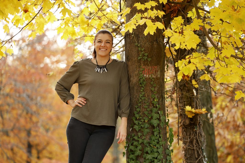 Lächelnde Frau lehnt an einem Baum mit gelben Herbstblättern.