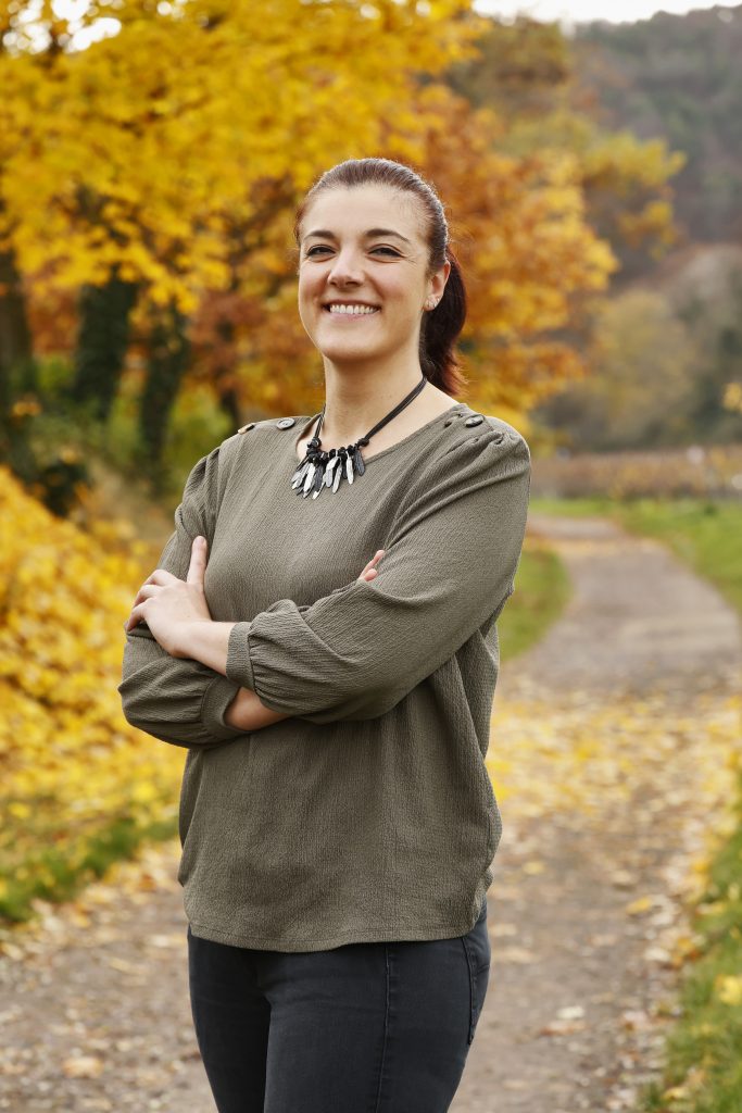 Lächelnde Frau steht mit verschränkten Armen auf einem Weg, umgeben von gelben Herbstbäumen.