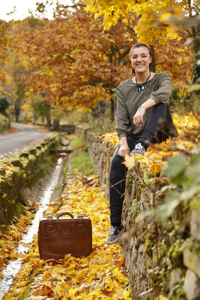 Lächelnde Frau sitzt auf einer Steinmauer am Wasserlauf, unten steht ein brauner Lederkoffer im Herbstlaub.