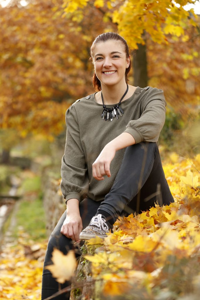 Lächelnde Frau sitzt auf einer Steinmauer, umgeben von gelbem Herbstlaub.