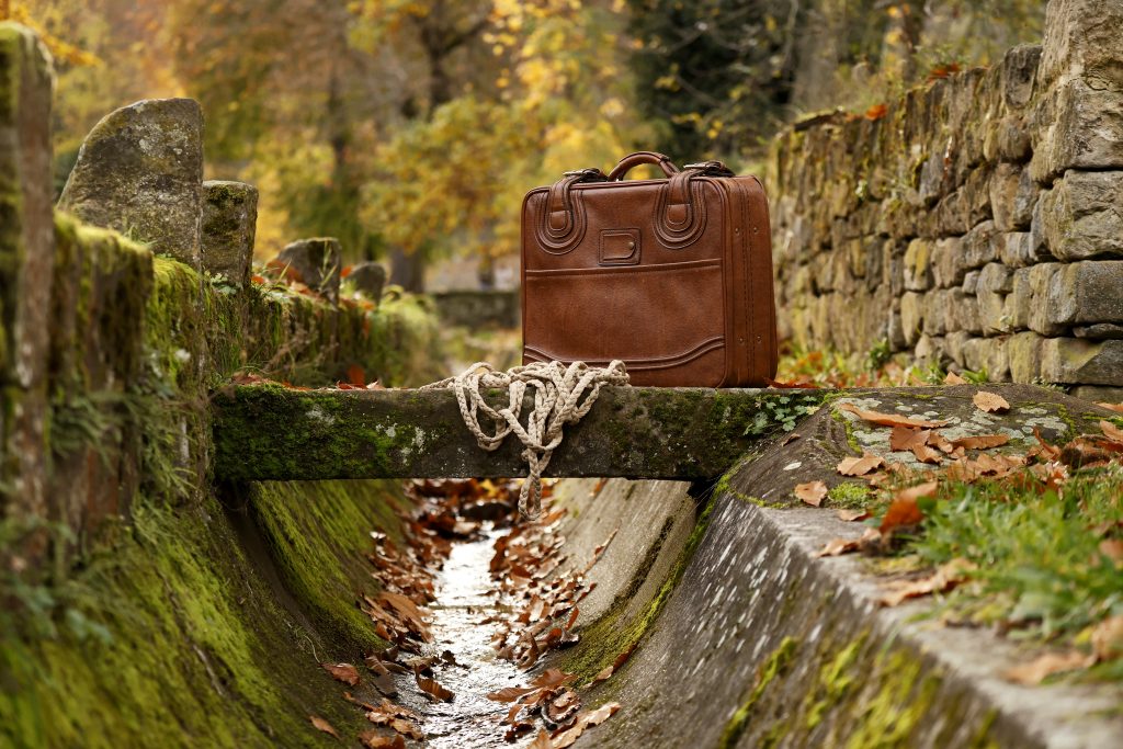 Brauner Lederkoffer steht auf einer Steinbrücke über einem schmalen Wasserlauf im Herbst.
